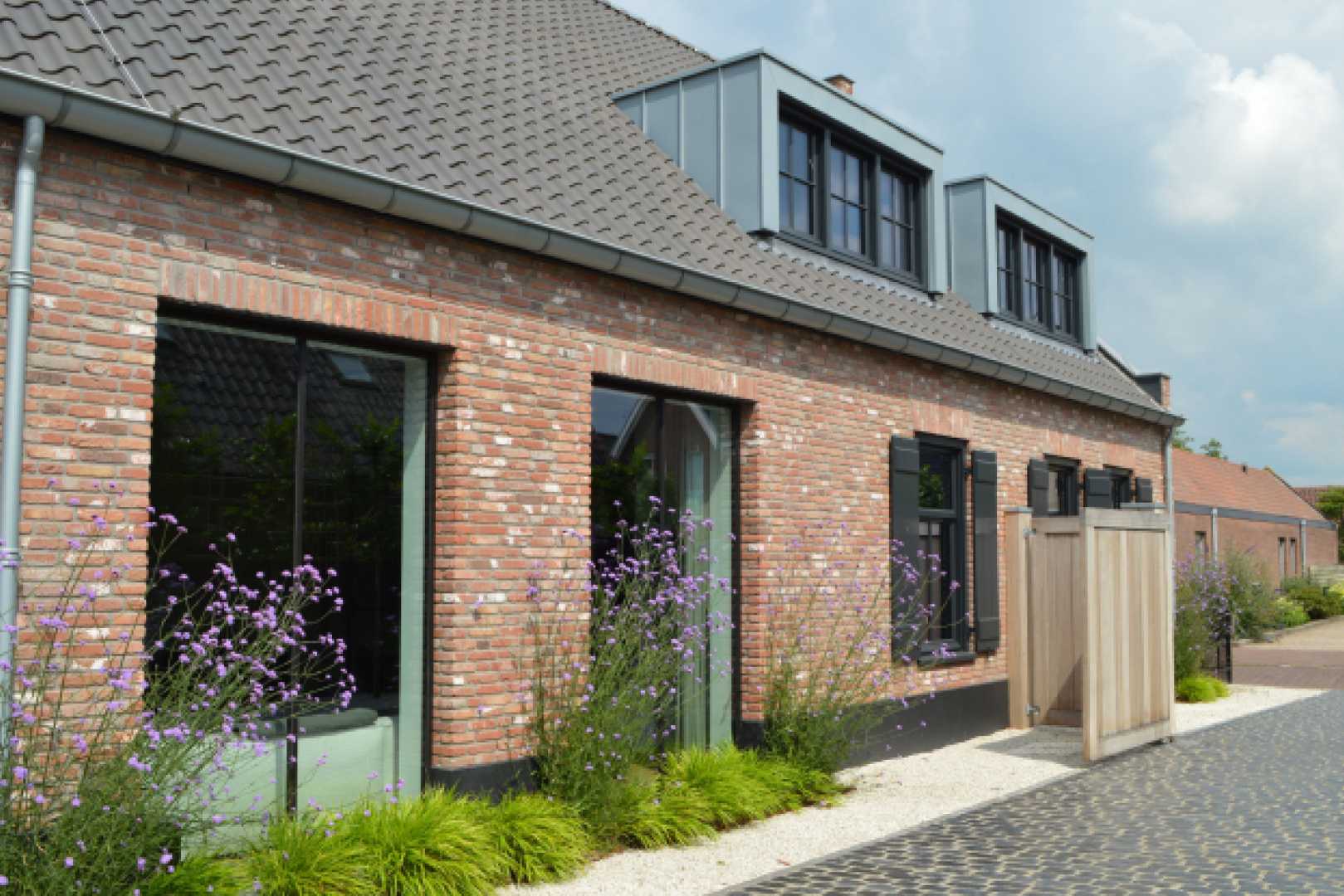 Vooraanzicht van bakstenen boerderij-stijl woning met mooie paarse bloemen. Op het dak zijn twee dakkapellen aangebracht. Ze zijn bekleed met RHEINZINK prePATINA ECO ZINC blue-grey.