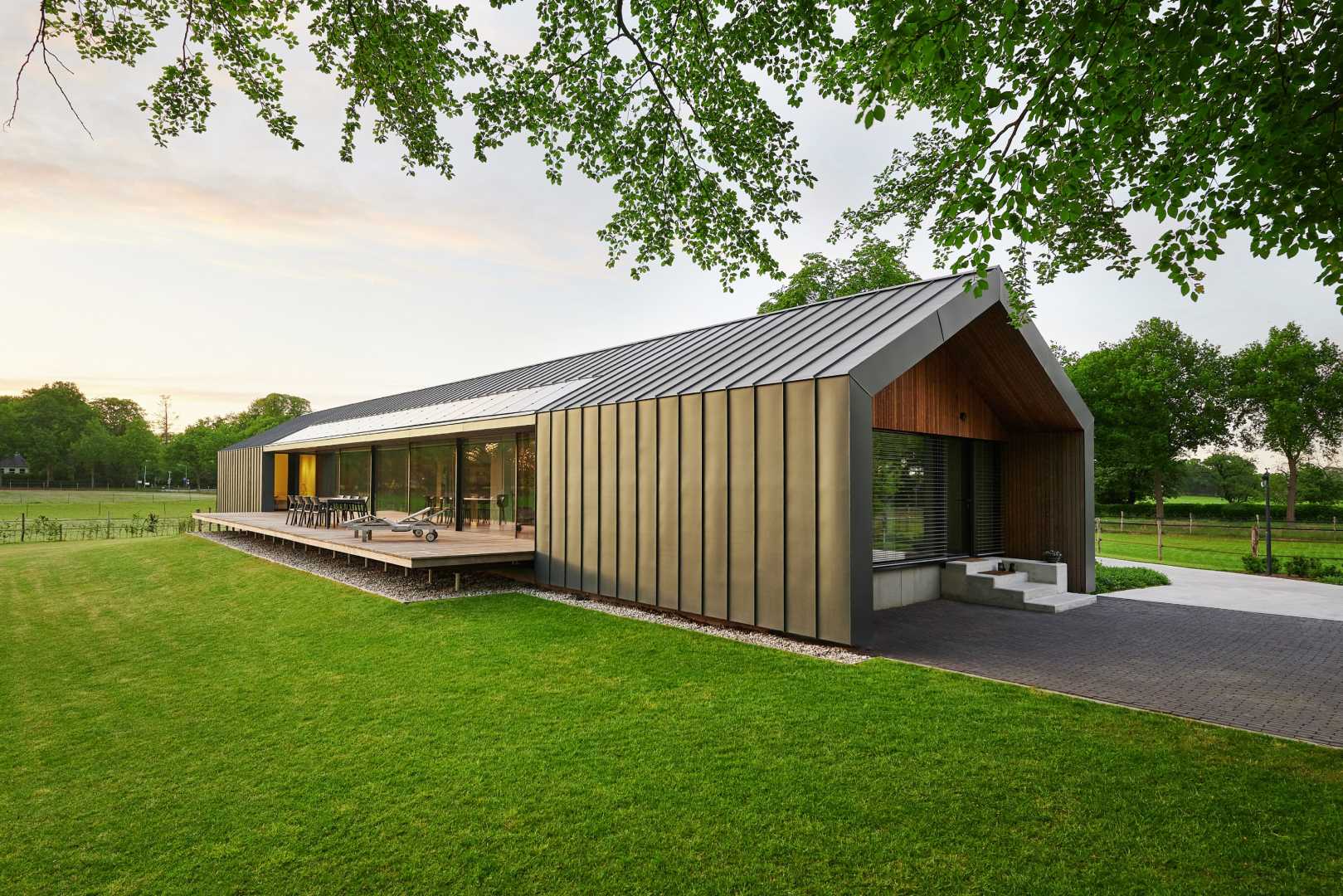 Moderne, langwerpige schuurwoning met terras gelegen in een groene weiland. De late middagzon weerkaatst zachtjes van de gevel en de daken die een harmonieus geheel vormen omdat ze allen bekleed zijn met RHEINZINK prePATINA ECO ZINC.
